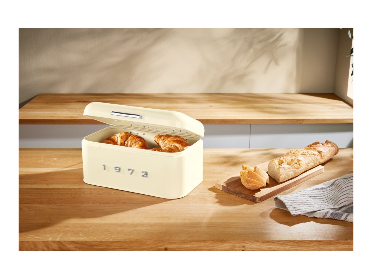 Retro-style cream bread bin with croissants inside, next to a baguette on a wooden counter.