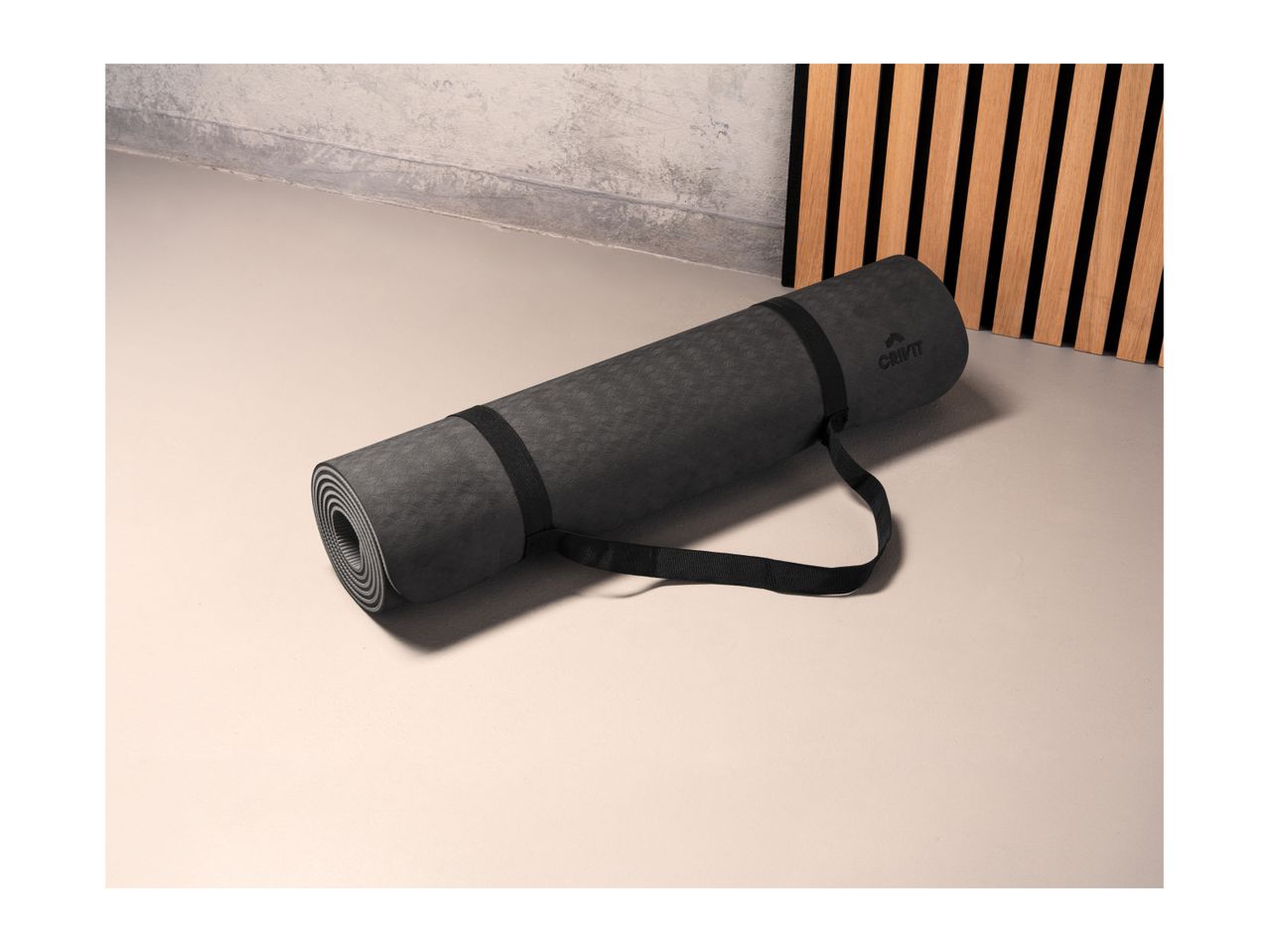 A rolled-up black Crivit yoga mat with a carrying strap on a light-colored floor.