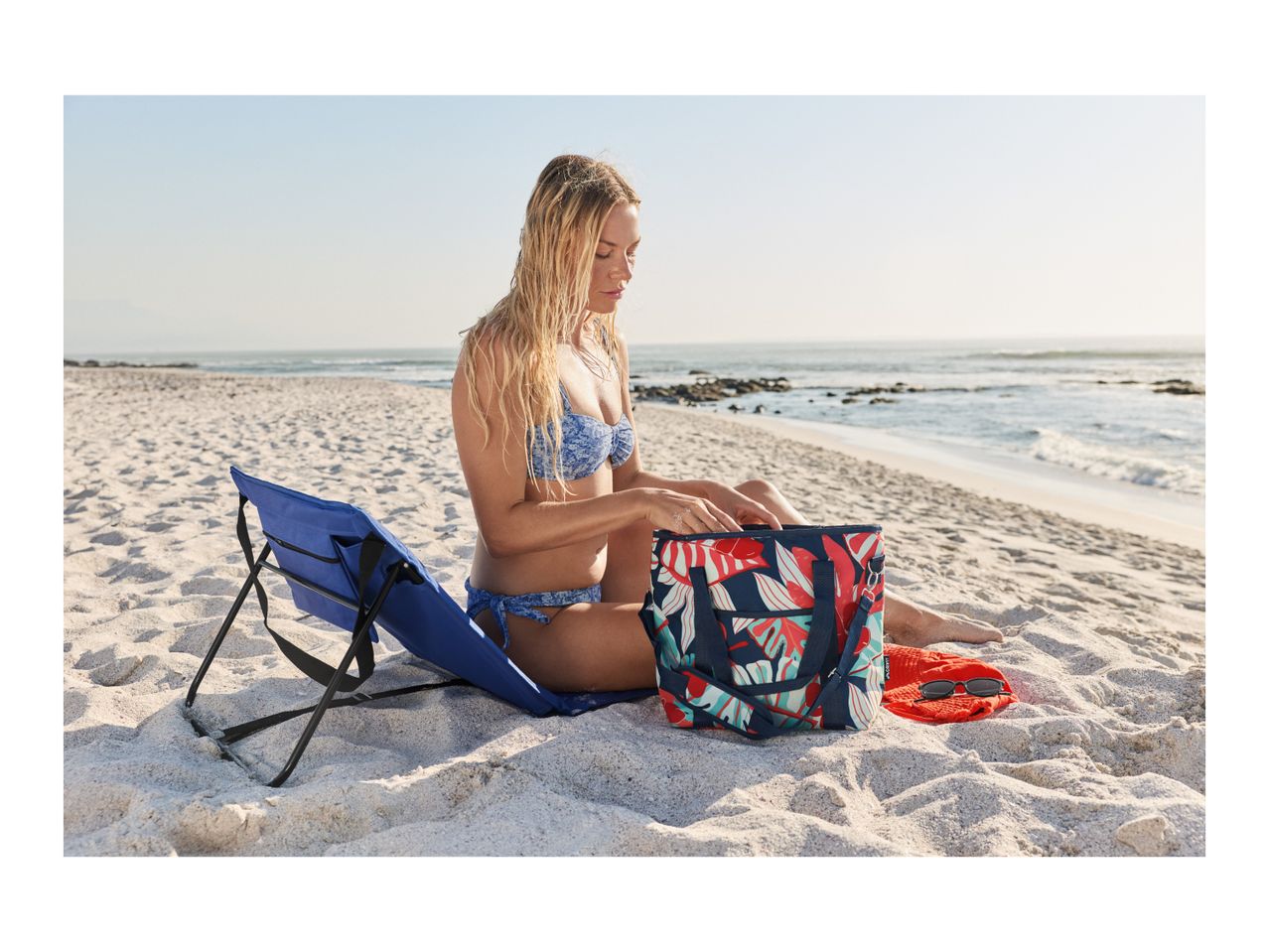 Woman in bikini on beach, sitting next to a blue beach chair and a floral cooler bag.
