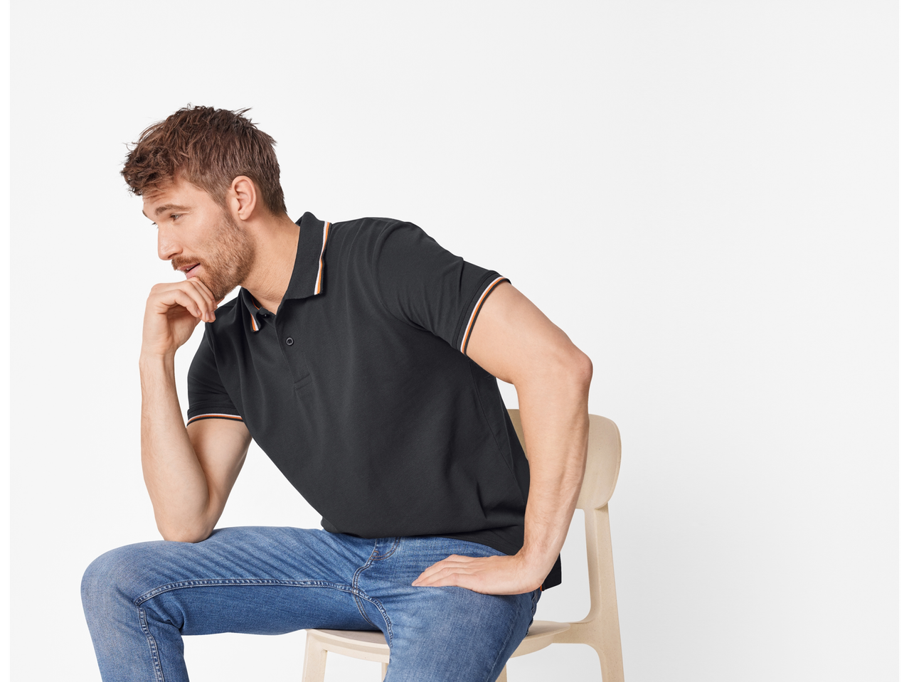 Un bărbat gânditor așezat pe un scaun, îmbrăcat cu un polo negru și blugi.