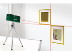 Nível a laser Parkside em tripé, projetando linhas vermelhas numa parede com dois quadros.