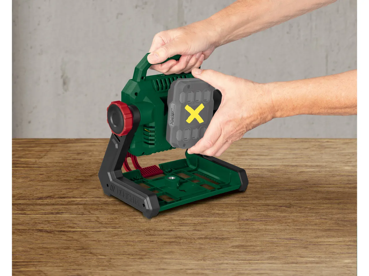 Mani che inseriscono una batteria grigia con una X gialla in un faretto da lavoro verde e nero.