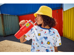Woman in a yellow bucket hat and grocery-themed shirt drinking from a red tumbler.