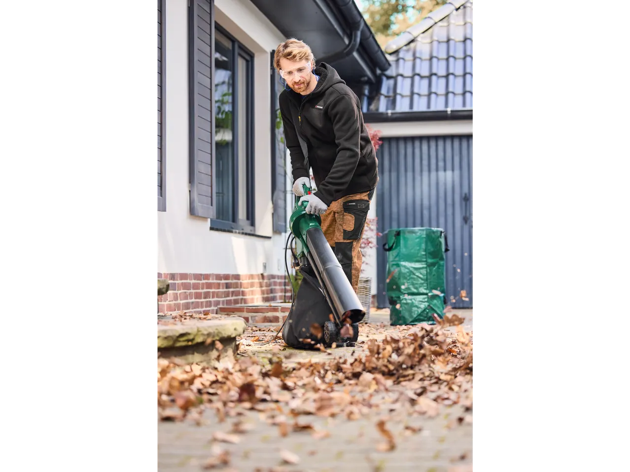 A man uses a Bosch leaf blower to clear autumn leaves from a patio.