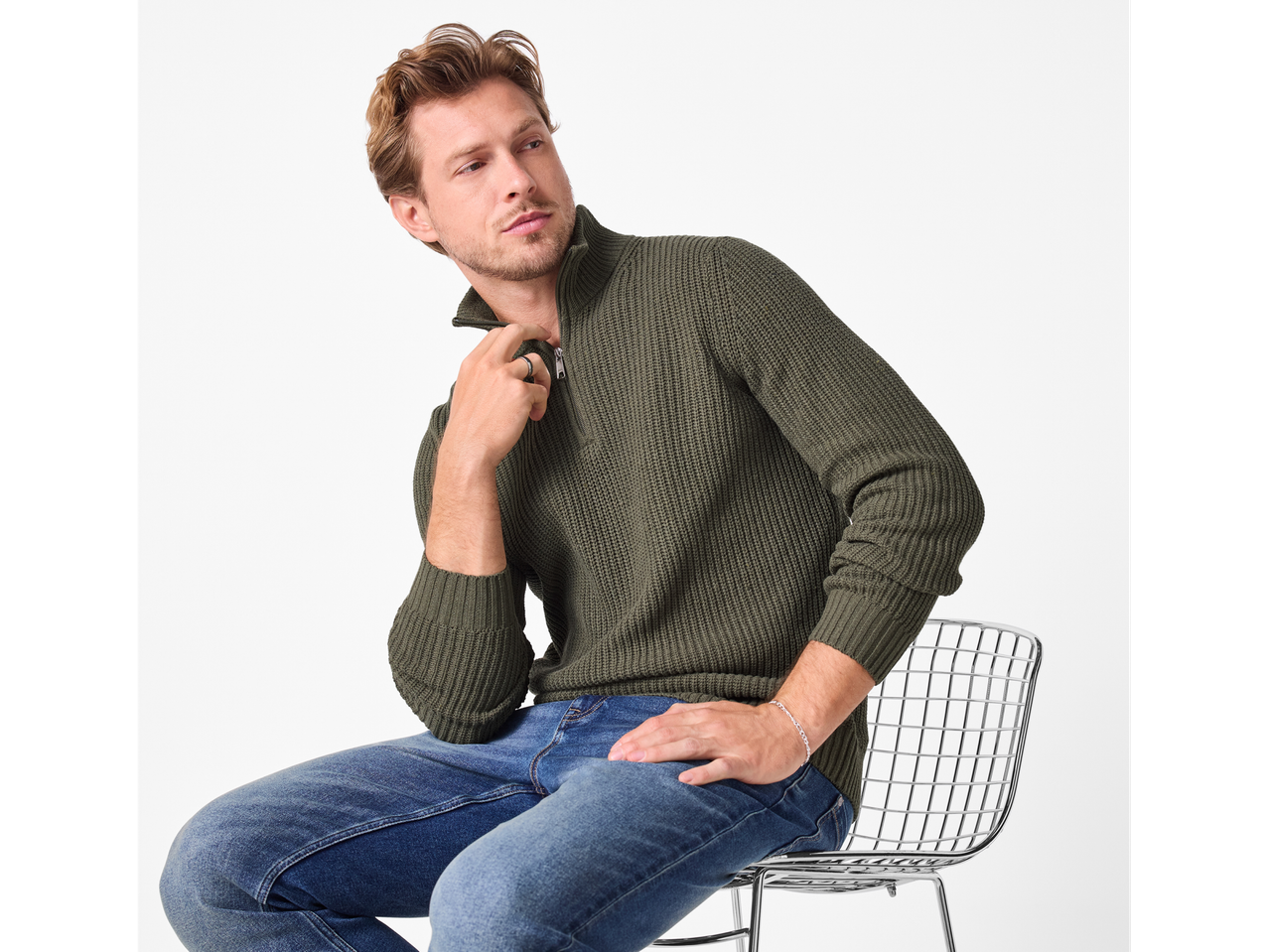 Vyras su žaliu Esmara Men megztiniu ir mėlynais džinsais, sėdi ant kėdės.