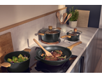 Black cookware set with wooden handles on a kitchen counter, cooking meat and vegetables.
