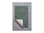 A green bath mat with a corner folded back, revealing its underside, on a gray tiled floor.
