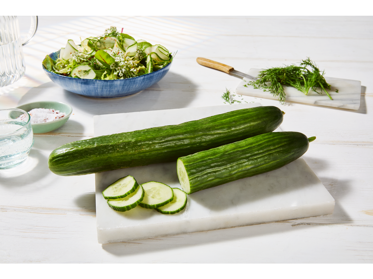 Two fresh cucumbers, one whole and one sliced, on a white marble board with a salad in the background.