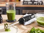 A black immersion blender sits on a kitchen counter next to a glass of green smoothie and a bowl of ingredients.