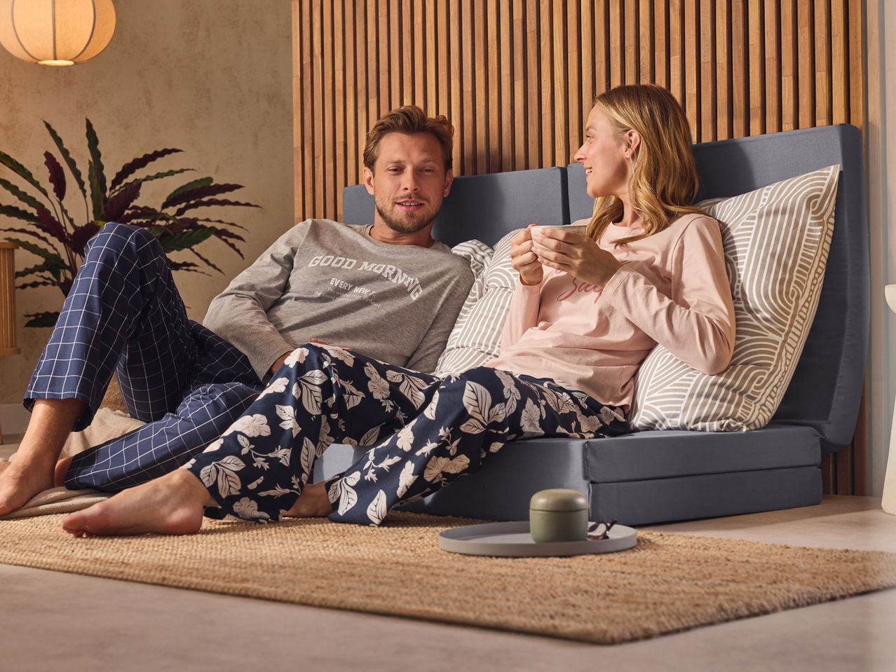 A couple in pajamas relaxing on a foldable mattress, drinking coffee.