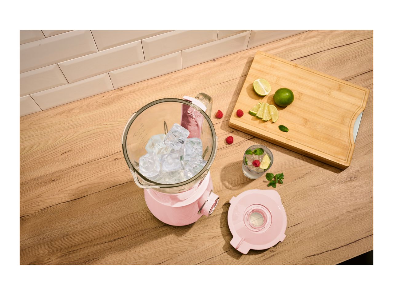 Pink blender with ice cubes, limes, raspberries, and a drink on a wooden counter.