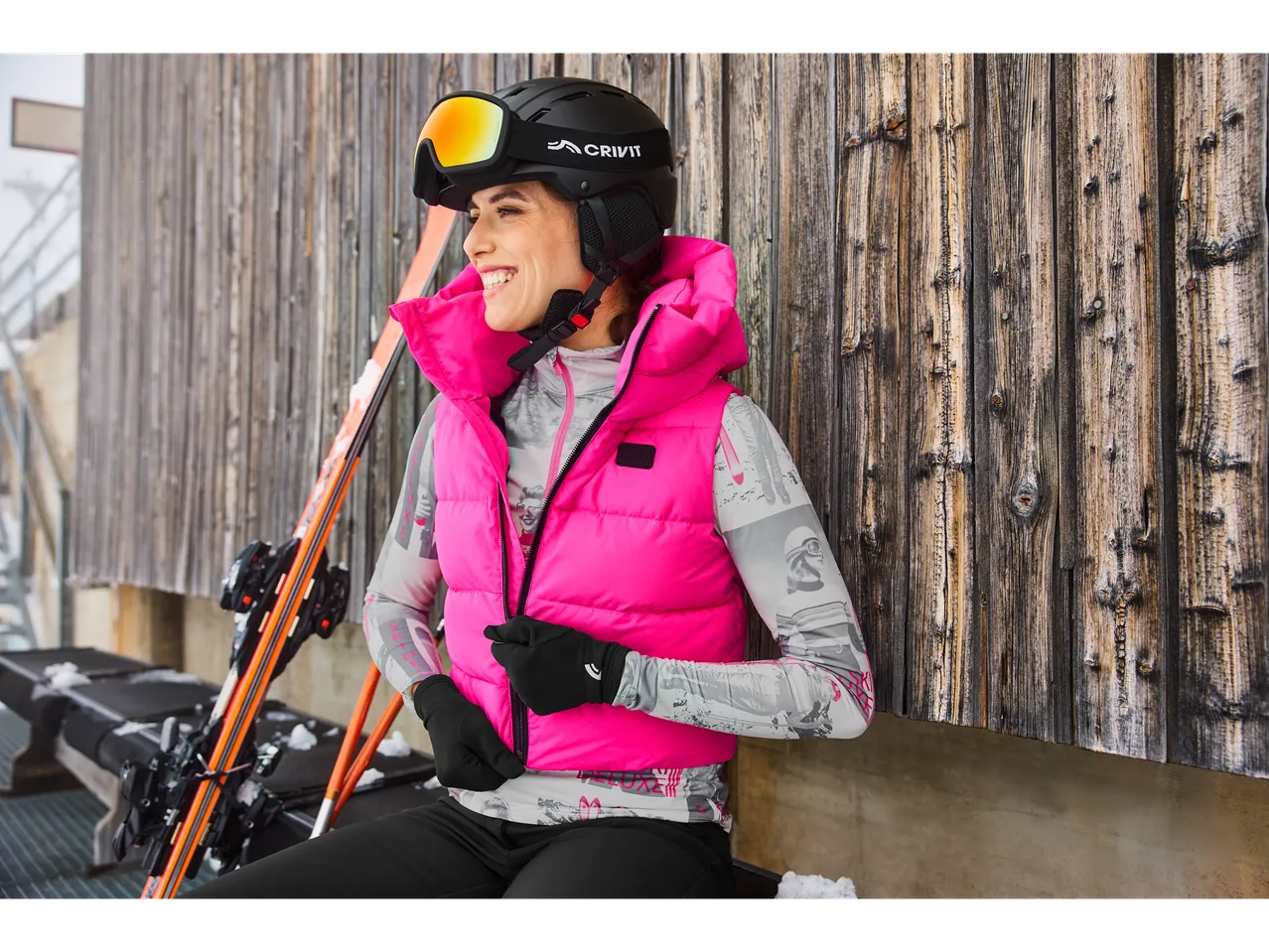 Woman in Esmara® ski base layer and pink vest, smiling by a wooden wall.