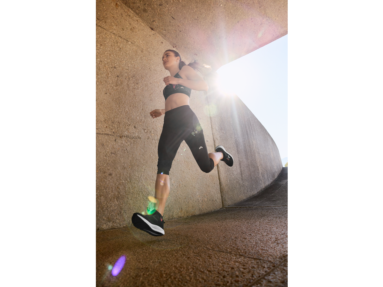 Woman in black sports bra and capri leggings running outdoors, sun flare in background.