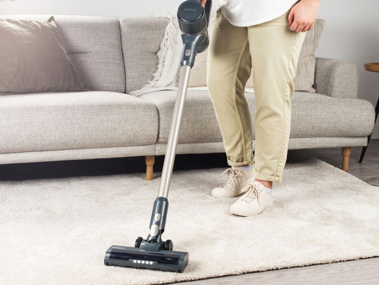 Person vacuuming a light-colored rug with a cordless stick vacuum cleaner.