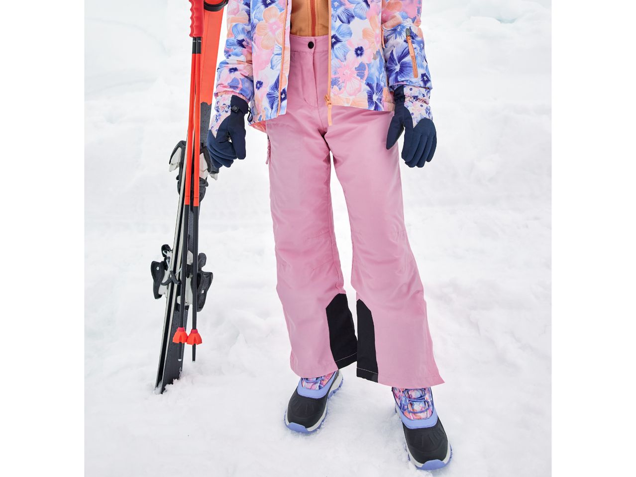 Person in pink ski pants and floral jacket, holding skis and poles in snow.