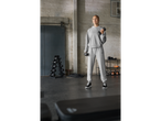 A woman in a grey tracksuit holds dumbbells in a gym with a dumbbell rack and kettlebells.