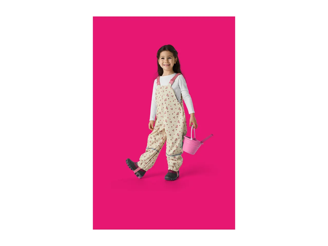 Smiling girl in floral overalls holding a pink watering can.