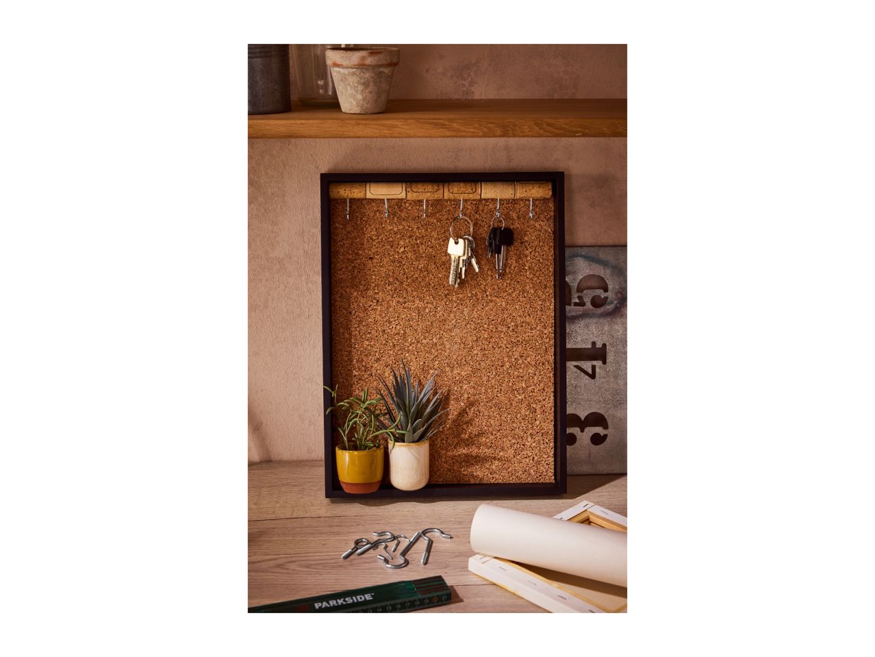 A cork board with keys hanging on hooks, two potted plants, and tools on a wooden surface.