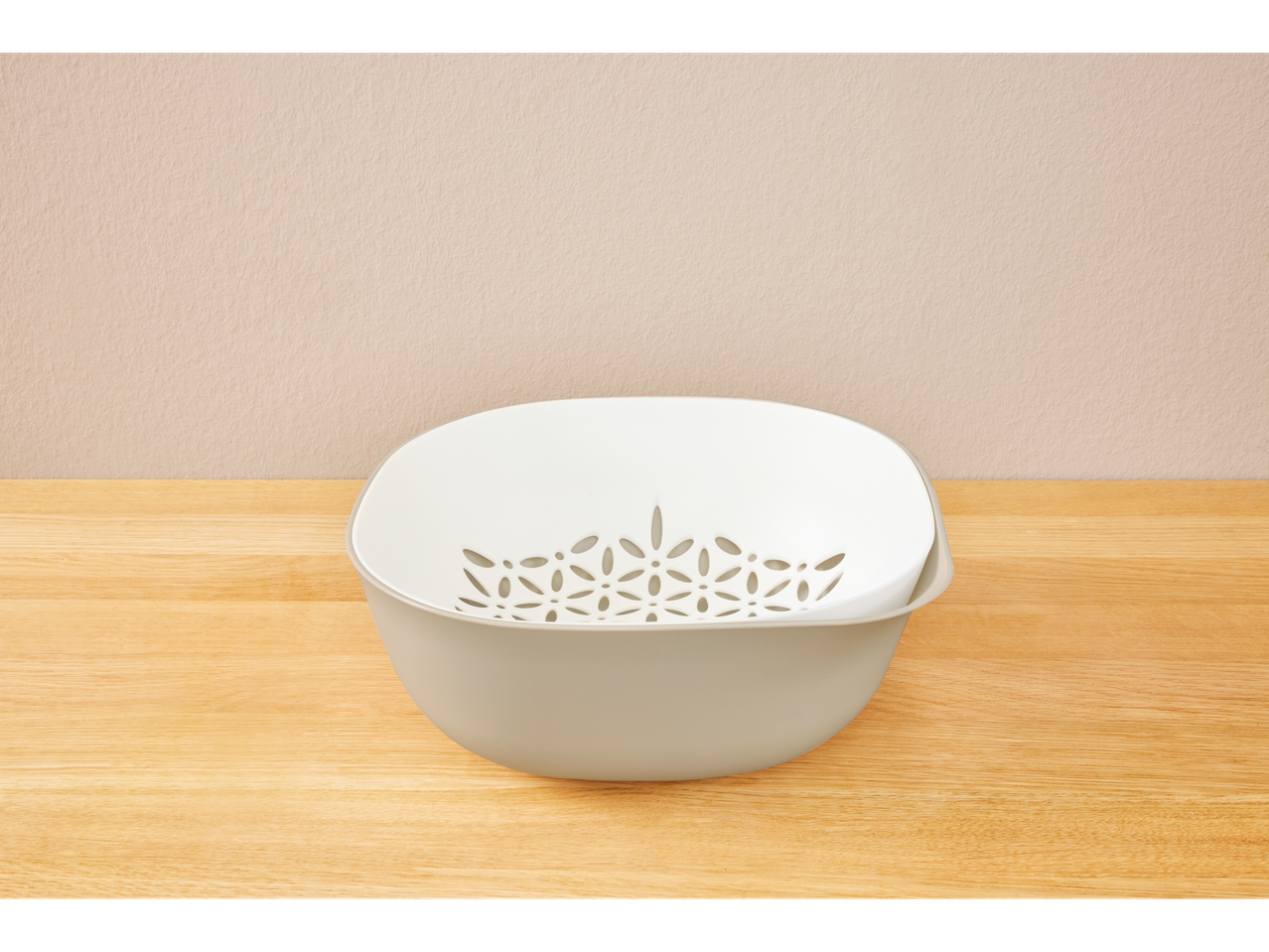 A beige plastic colander with a white inner bowl sits on a light wood surface.