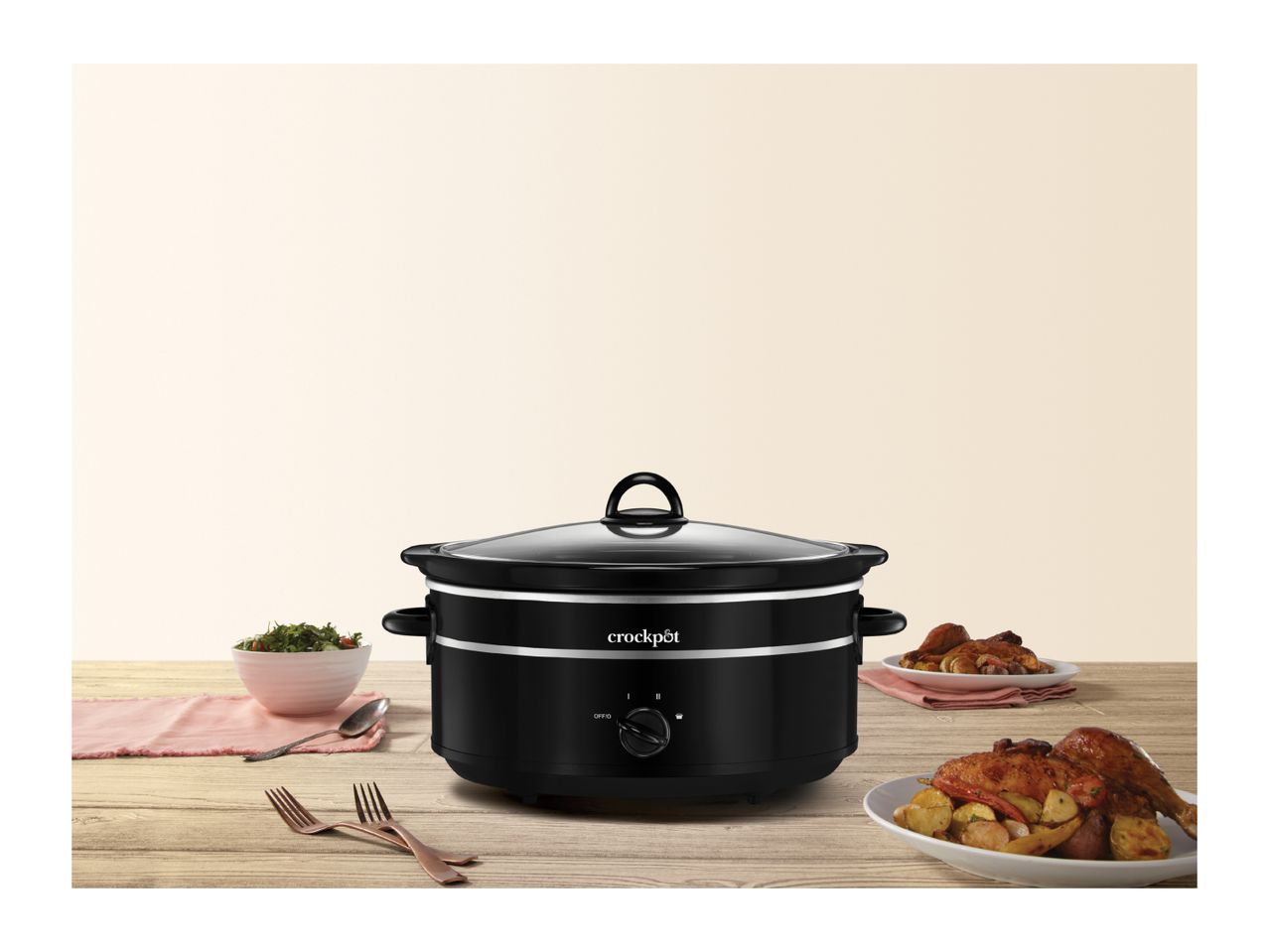 Black Crock-Pot slow cooker on a wooden table with salad and roasted chicken.