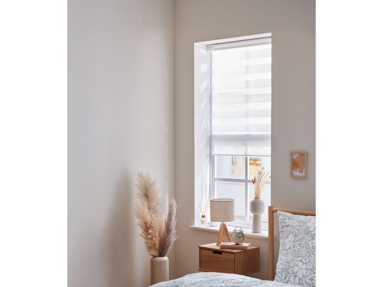 A bedroom with a wooden bedside table, lamp, alarm clock, and pampas grass in a vase.
