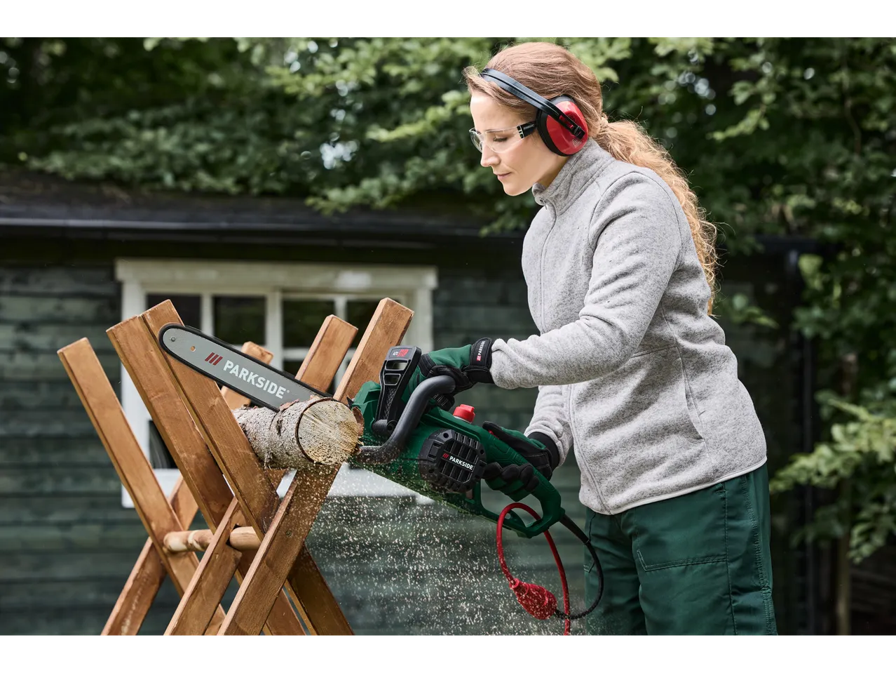 Femeie tăind un buștean cu un fierăstrău electric, purtând protecție pentru ochi și urechi.