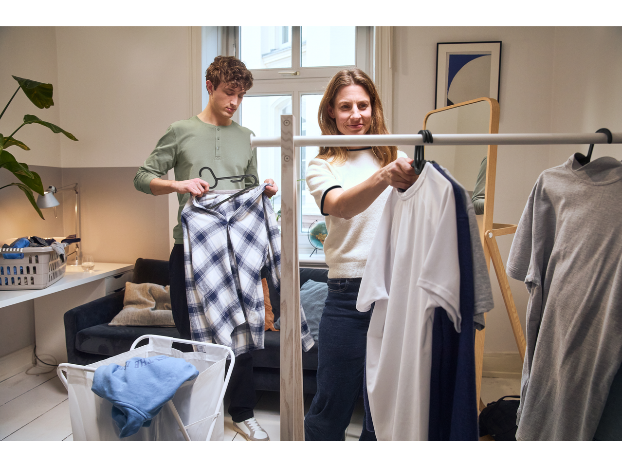 Two people hanging clothes on a rack, with laundry baskets and a desk in the background.