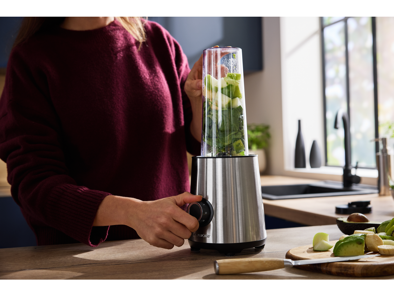 Mulher a preparar um smoothie com o liquidificador SILVERCREST, adicionando frutas e vegetais.