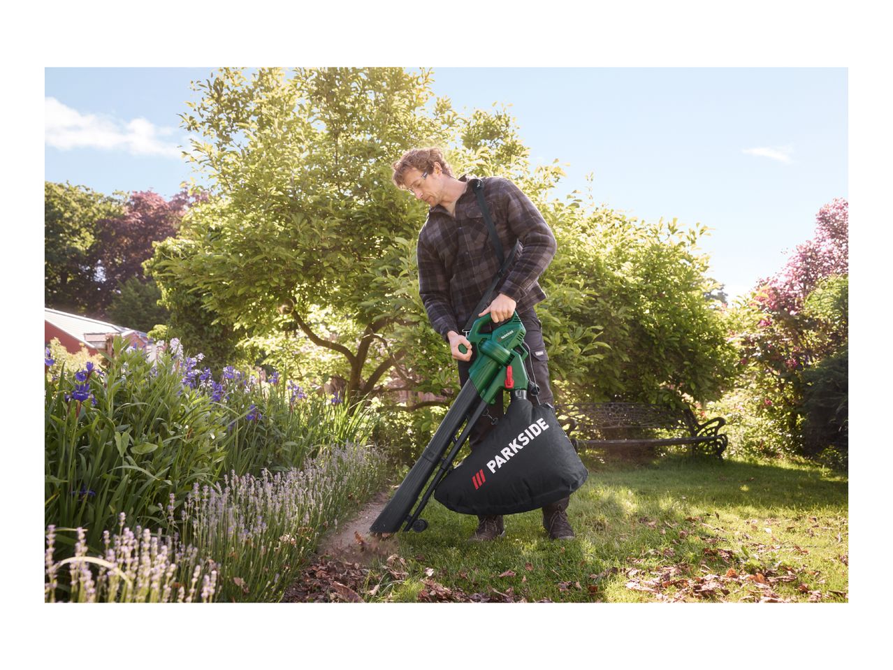 Man using a leaf blower/vacuum in a garden, clearing leaves from a flower bed.