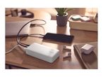 White power bank charging a smartphone and laptop on a wooden desk, with earbuds nearby.
