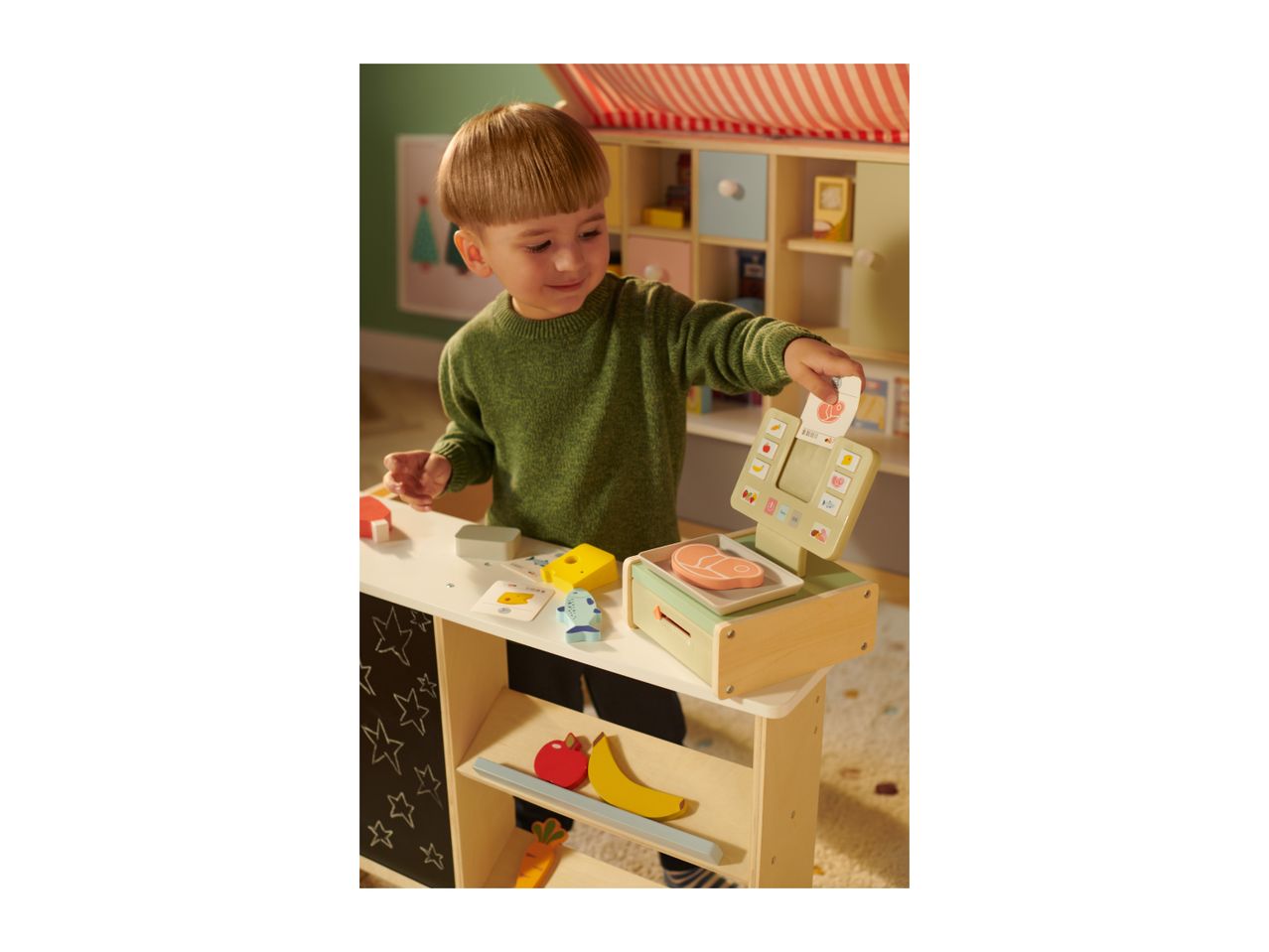 A child playing with a wooden toy market stall, featuring a scale, food items, and a chalkboard.
