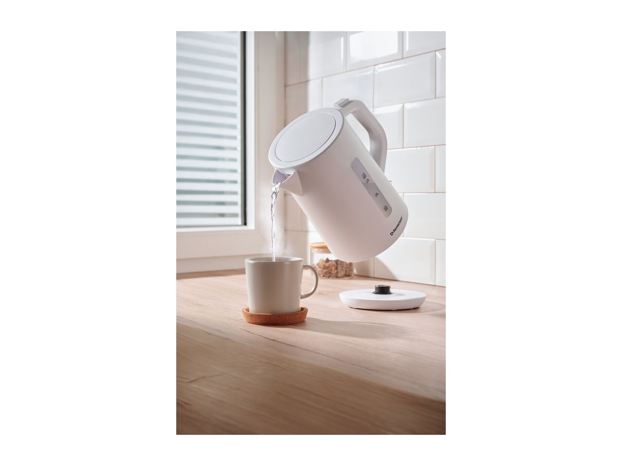 White electric kettle pouring hot water into a mug on a wooden countertop.