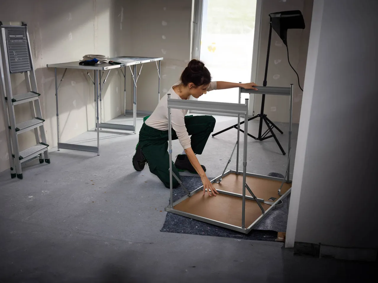 A woman assembling a work table in a room with a ladder and another table.