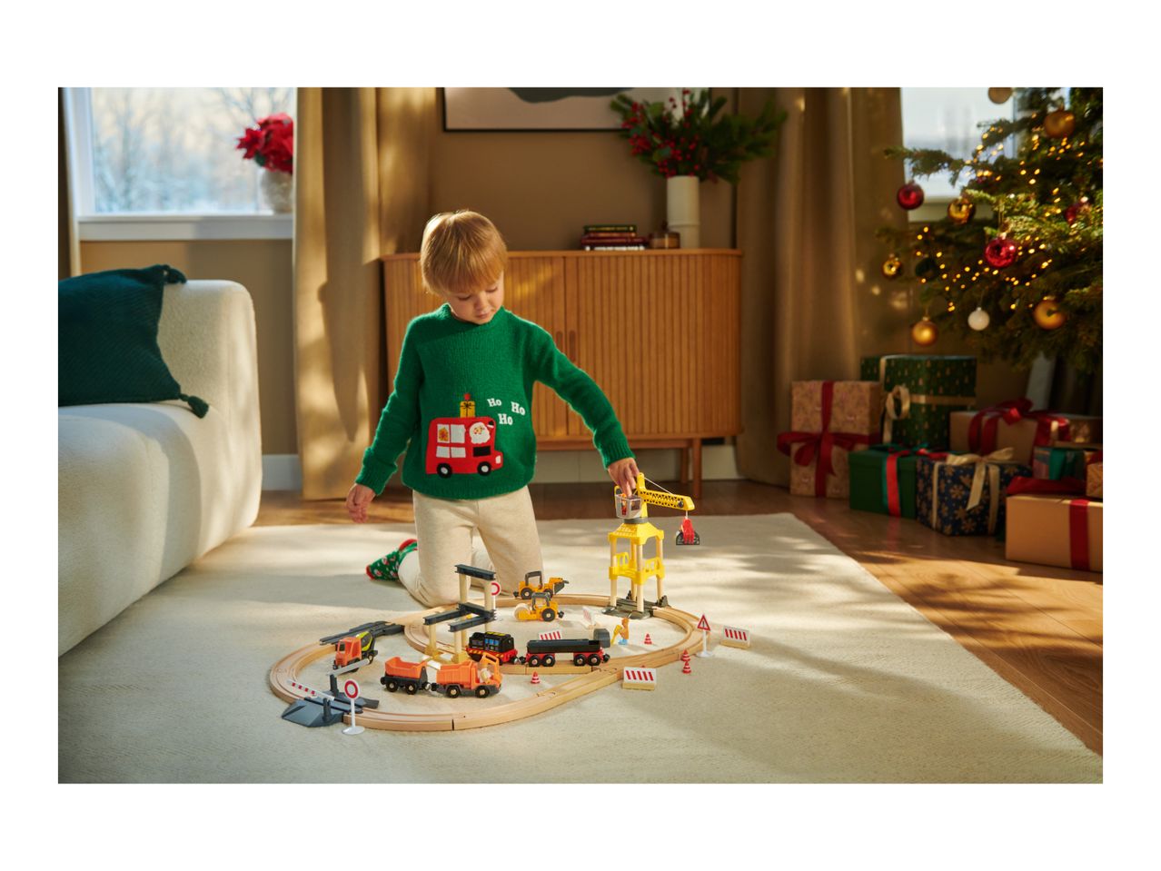 Child in Christmas sweater playing with a wooden train set and construction vehicles.