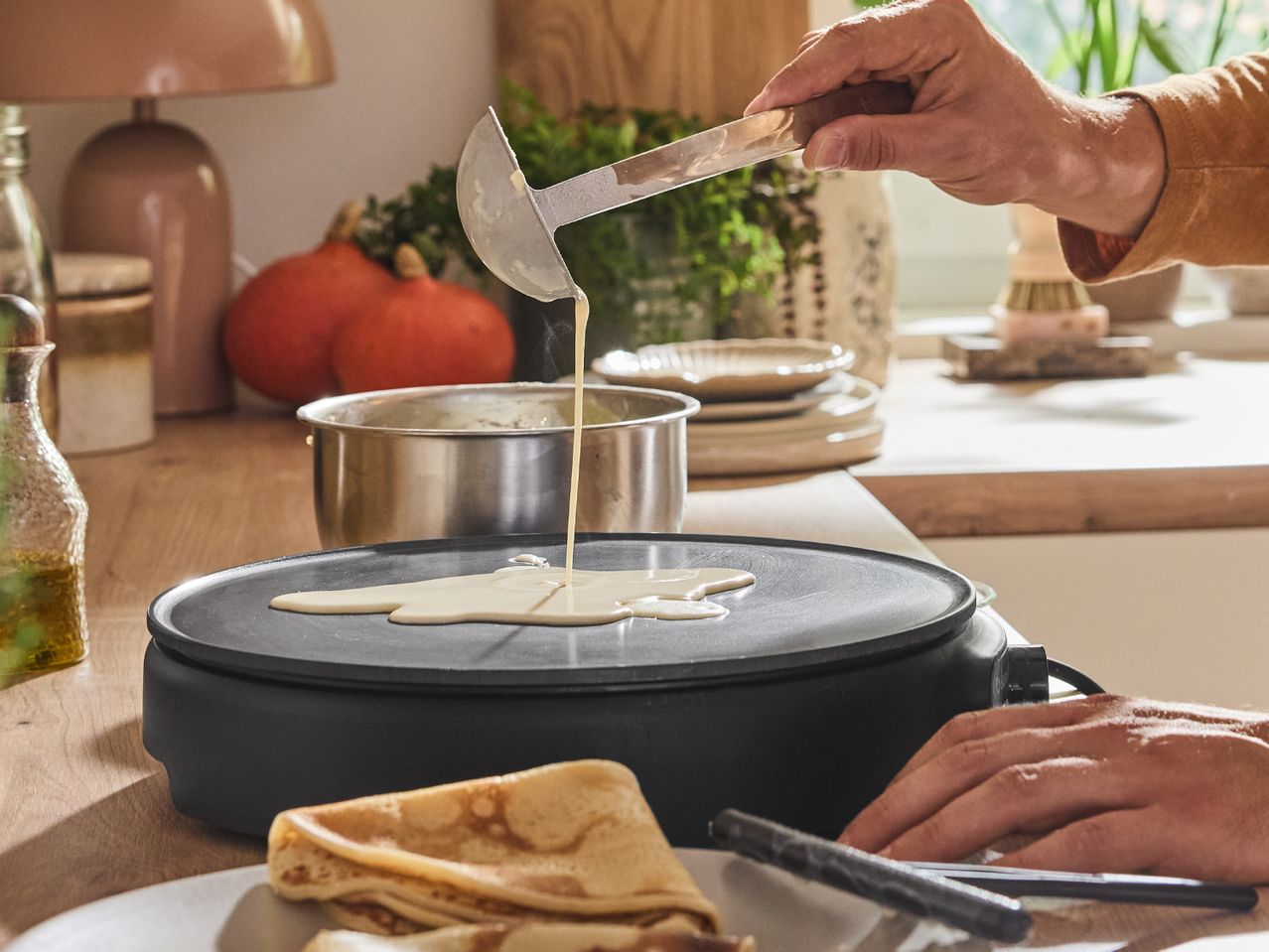 A person pouring pancake batter onto a crepe maker on a wooden kitchen counter.
