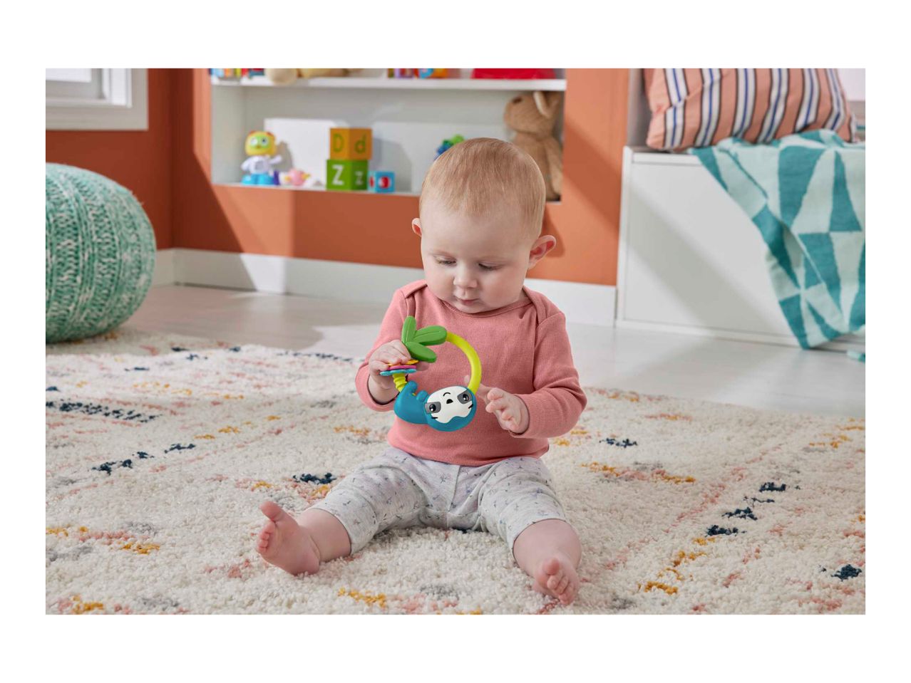 Baby playing with a sloth-themed rattle on a patterned rug in a nursery.