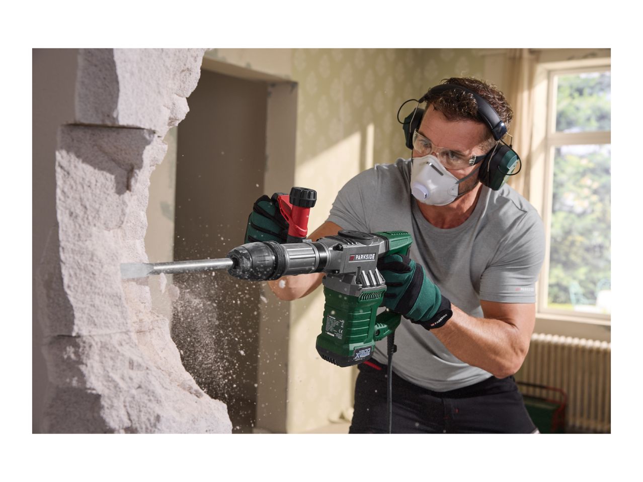 Man using a demolition hammer to break a wall, wearing safety gear.