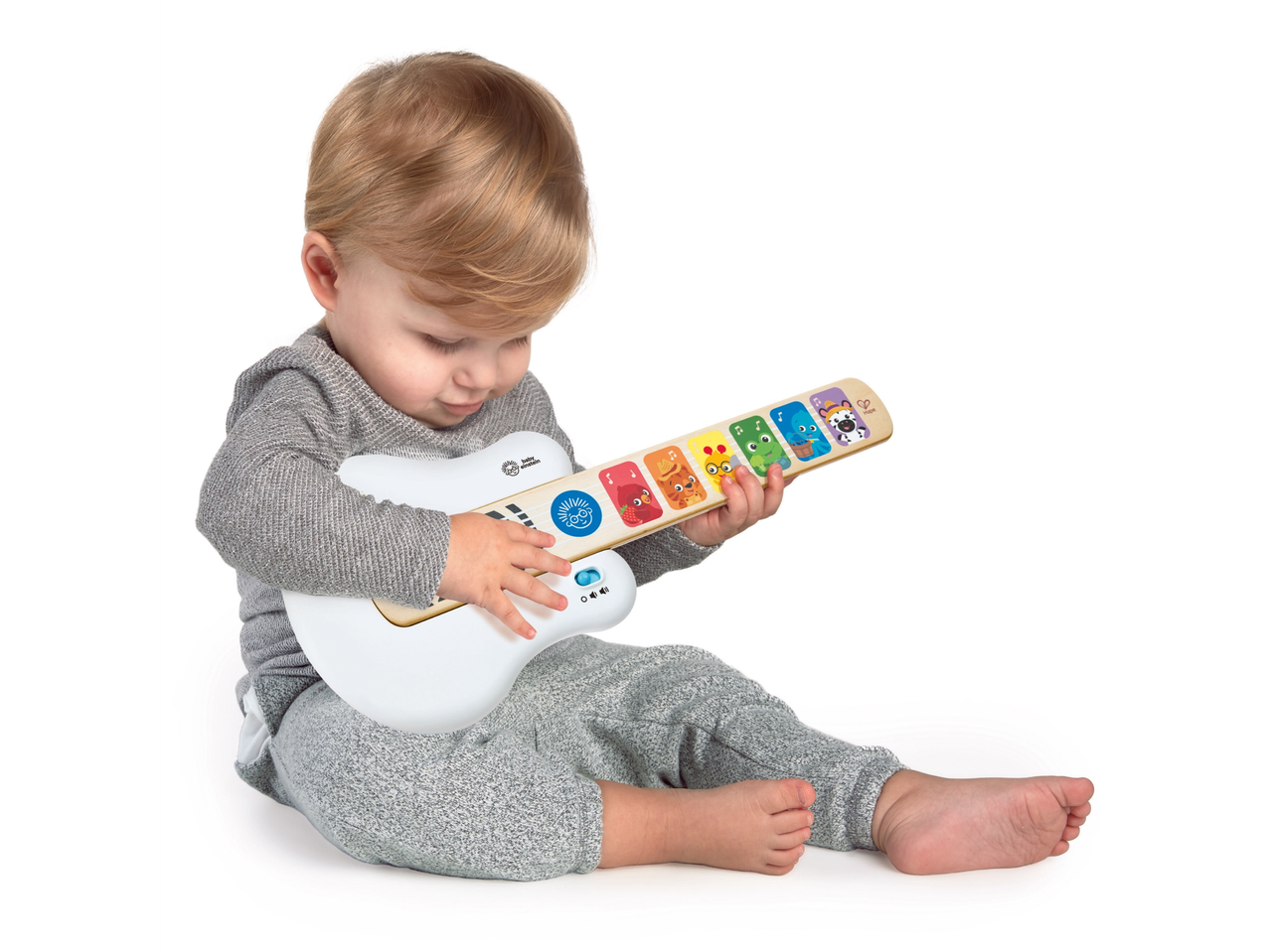 A toddler in a gray outfit sits on a white floor, playing a white and wooden Baby Einstein toy guitar.