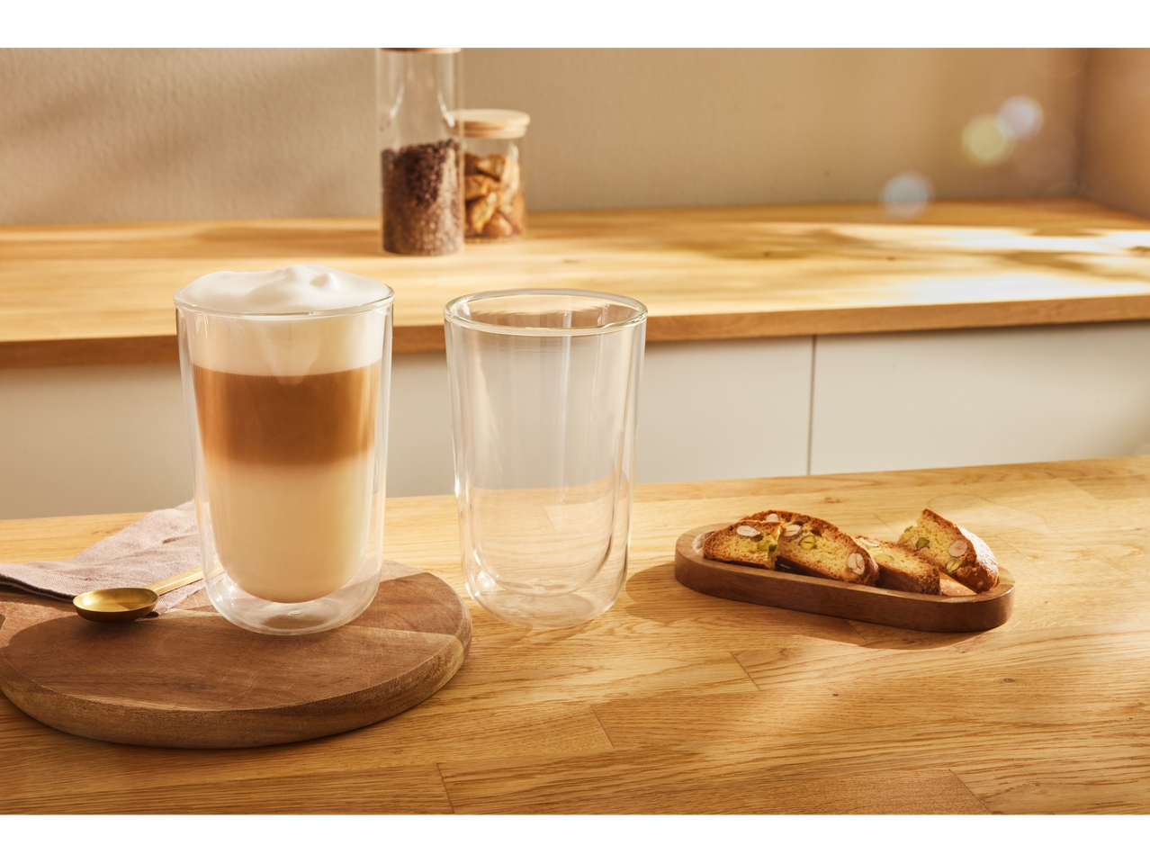 Um copo de café com leite e um copo vazio em uma mesa de madeira, com biscoitos ao lado.