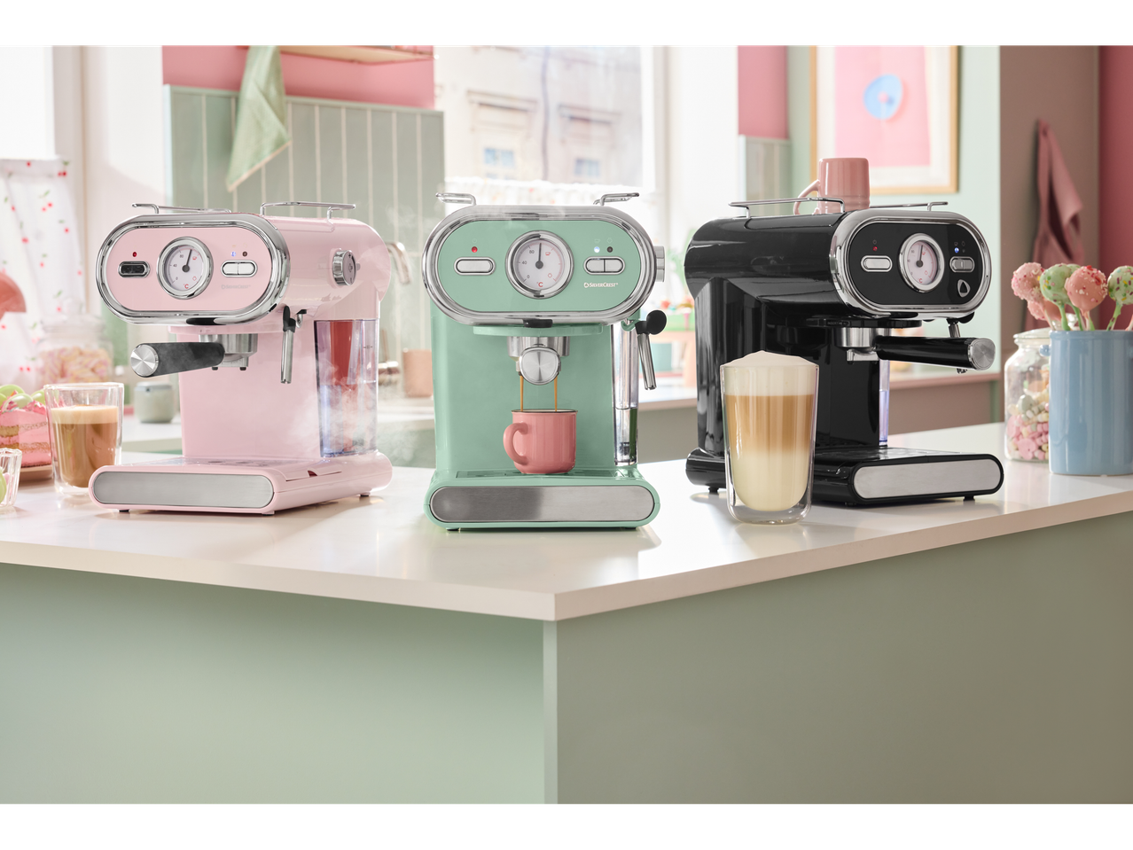 Three SilverCrest espresso machines on a kitchen counter: pink, mint green, and black.