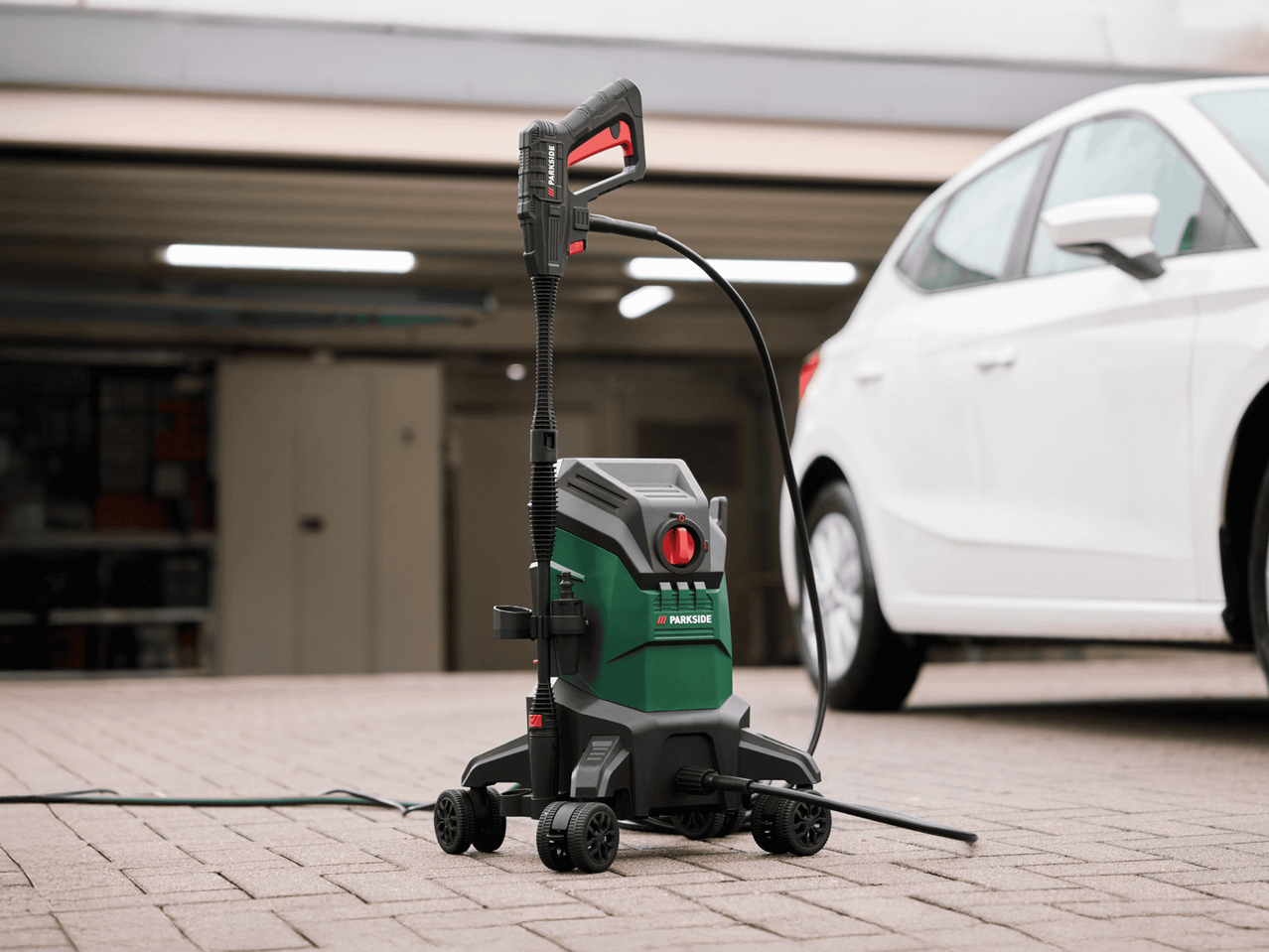 Hochdruckreiniger mit Sprühlanze und Schlauch vor einem weißen Auto auf Pflastersteinen.
