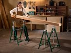 Woman in a workshop placing a wooden beam on two green saw horses.