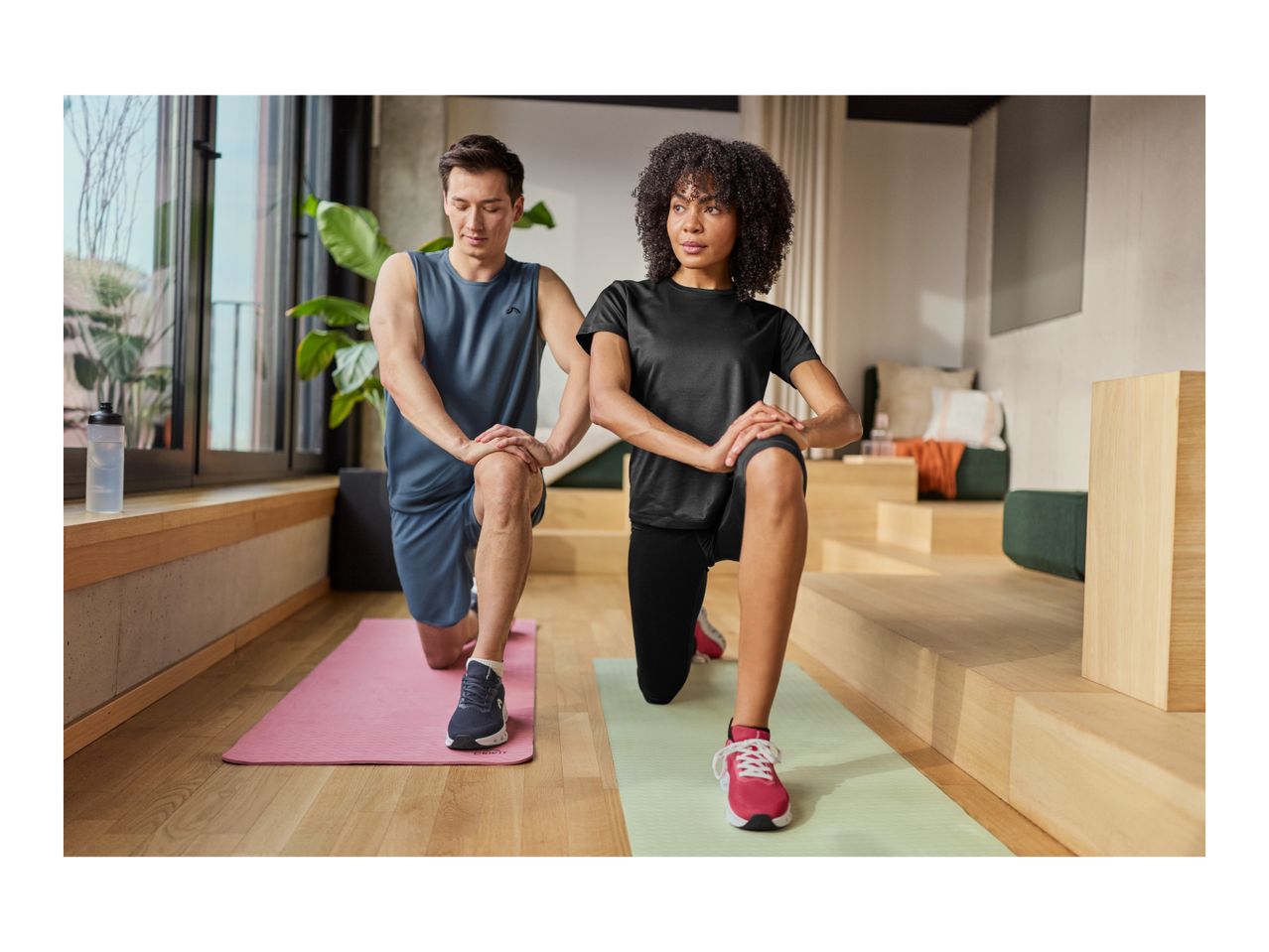 Man and woman in sportswear doing lunges on yoga mats in a modern living room.