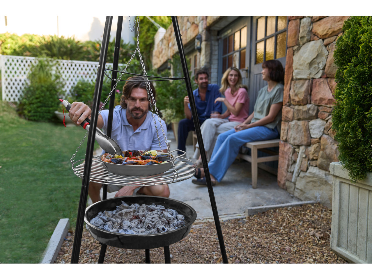 Grillmeister Iron Grill Pan with seafood over hot coals, friends relaxing in the background.