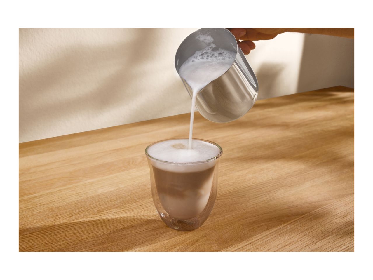 A hand pouring frothed milk from a metal pitcher into a glass of coffee on a wooden table.