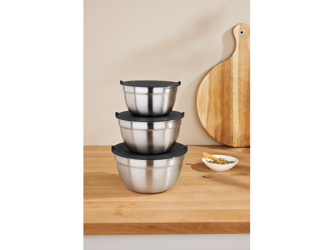 Stacked stainless steel mixing bowls with black lids on a wooden counter with a cutting board.