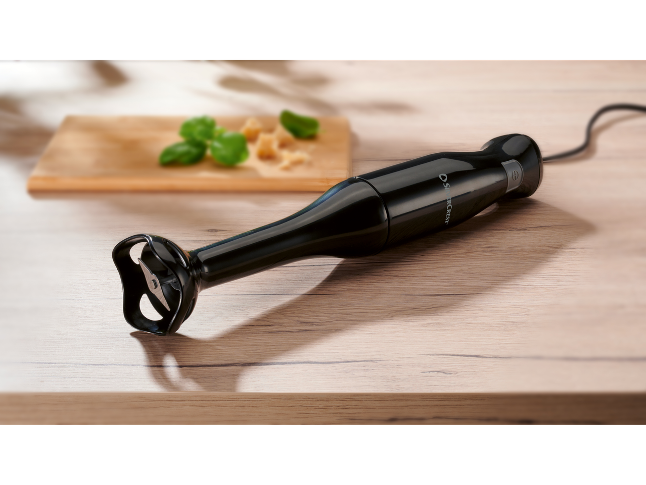 A black Silvercrest hand blender rests on a light wood surface near a cutting board with basil and cheese.