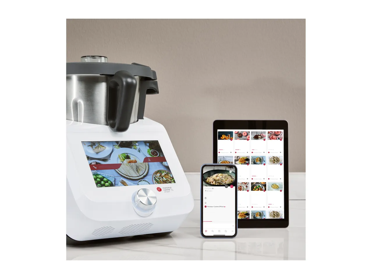 A white Monsieur Cuisine Smart food processor with a screen displaying recipes, next to a tablet and smartphone showing recipe apps.