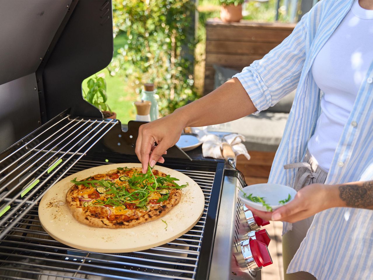Nő pizzát tesz egy grillen lévő pizzakőre, friss rukkolával díszítve.