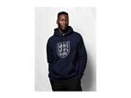Man in a navy blue hoodie with a white shield emblem, black trousers, hands in pockets.